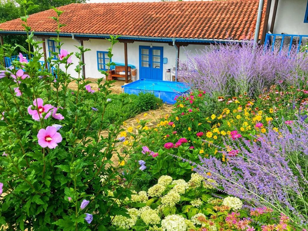 Property photo 1 of 64 View Of A Garden Filled With Various Colorful Flowers And A Small Blue Pool, With A White House Featuring Blue Doors And Windows And A Red Tiled Roof In The Background.