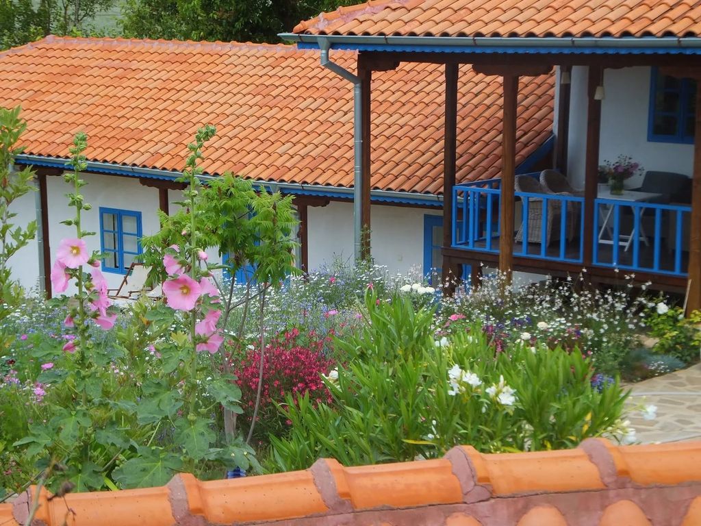 Property photo 2 of 64 A Garden With A Variety Of Flowers In Front Of A White House With Blue Window Frames And A Covered Porch Furnished With Wicker Chairs And A Small Table.
