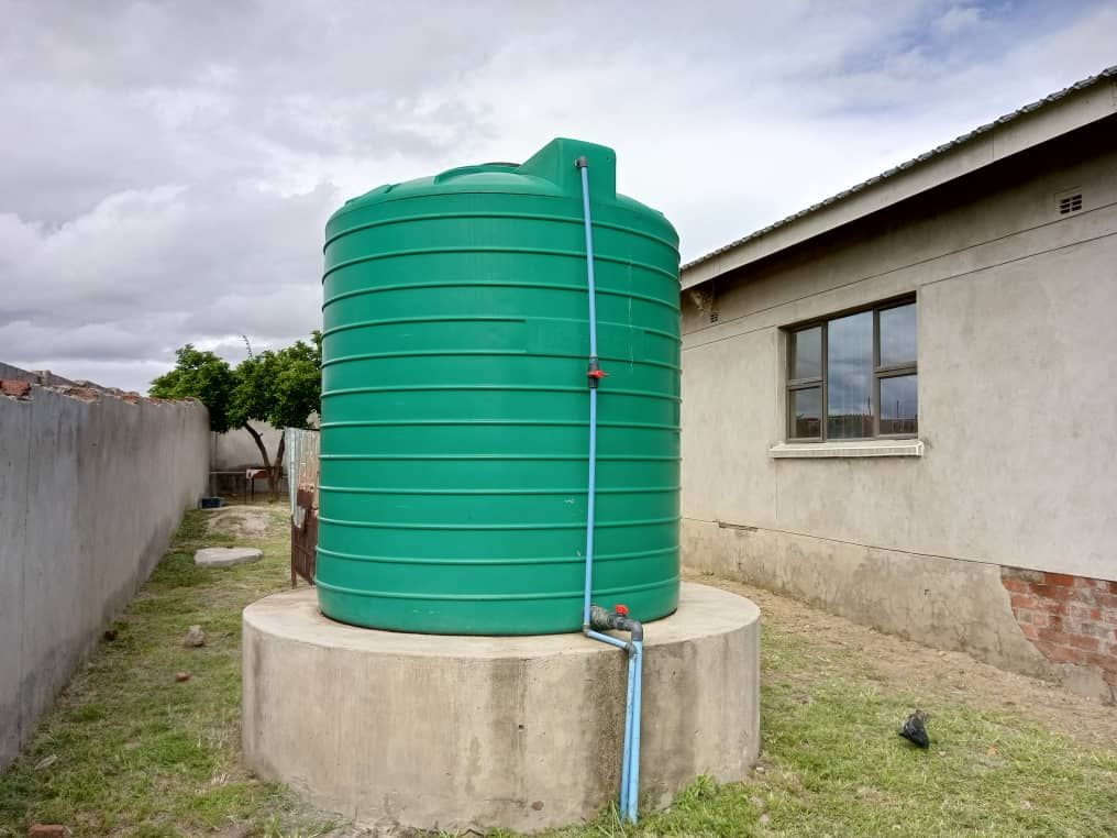 Property photo 3 of 9 Green Vertical Water Storage Tank Mounted On A Concrete Circular Base Beside A House, With Blue Piping And A Red Valve Line Running Up The Side.