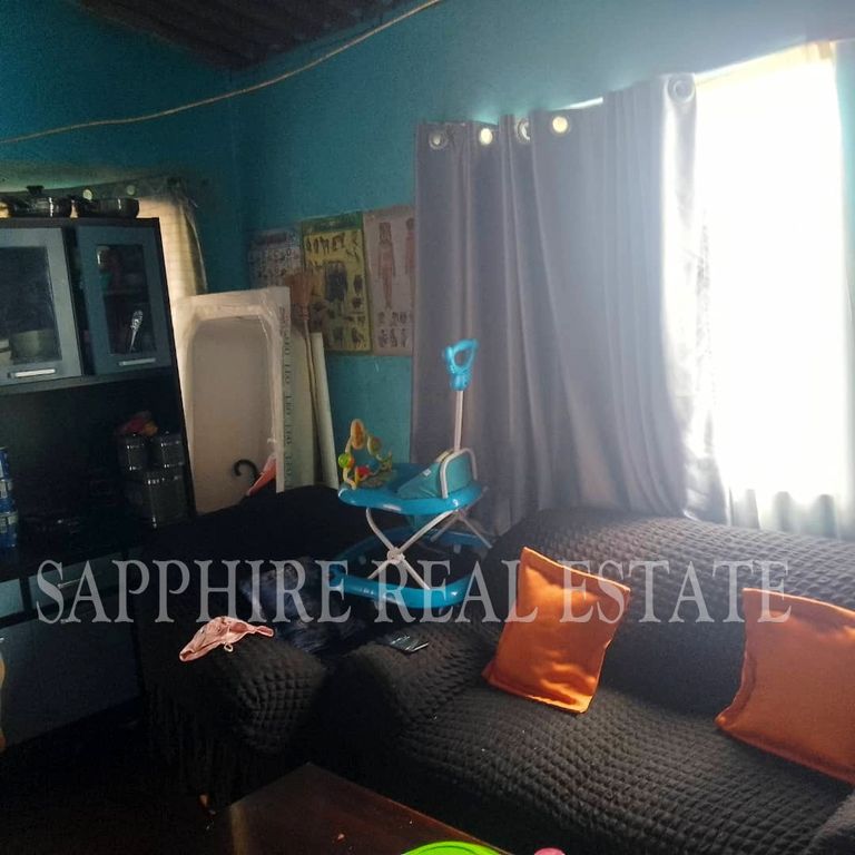Property photo 3 of 18 Living Room With Teal Walls, A Dark Textured Sofa With Orange Cushions, And A Blue Baby Walker Resting On The Couch Corner Near A Window Curtain.