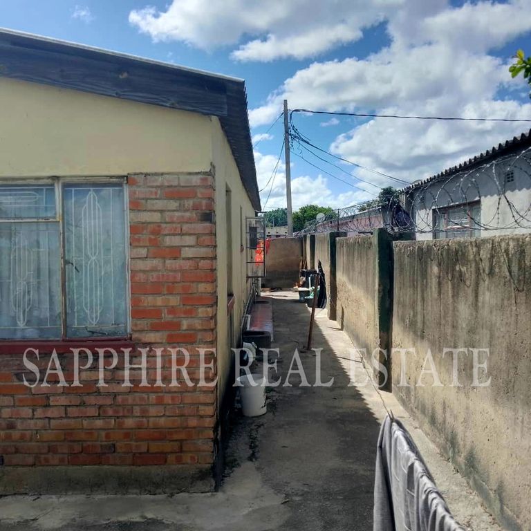 Property photo 2 of 18 Narrow Outdoor Alley Between A Brick House And A High Concrete Wall, With A Blue Sky And Barbed Wire Along The Top Of The Wall.