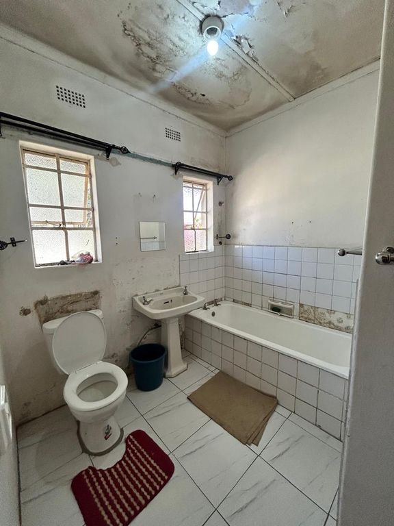 Property photo 3 of 9 Small Worn Bathroom With Toilet, Pedestal Sink, And Bath; Peeling Ceiling, Bare Bulb, Two Small Windows, And A Blue Bucket Nearby.