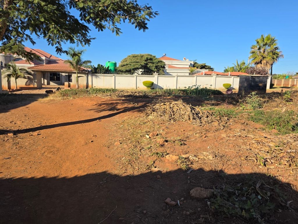 Property photo 1 of 5 Empty Dirt Lot In A Suburban Yard With A White Boundary Wall, Red-Tiled Houses, And Trees Under A Bright Blue Sky.