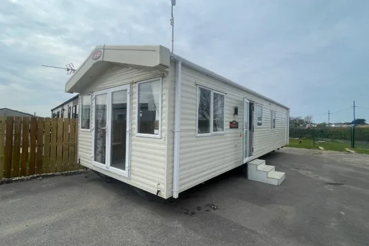 Property photo 1 of 10 A White Caravan Lodge With Windows And Doors On A Paved Lot Near A Wooden Fence Under A Cloudy Sky