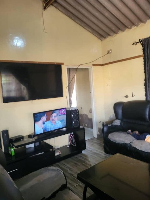 Property photo 3 of 7 Living Room With A Wall-Mounted TV, A Smaller TV On A Black Entertainment Unit, And A Person Resting On A Black Leather Sofa.