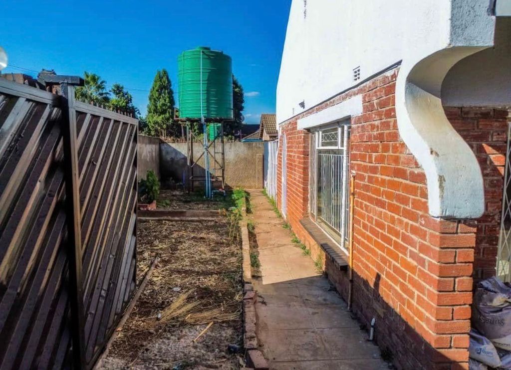 Property photo 3 of 6 Backyard Path Along A Brick House With A Tall Green Elevated Water Tank On Metal Supports, A Leaning Wooden Fence, And Dry, Bare Ground On The Left Side Under A Clear Blue Sky.