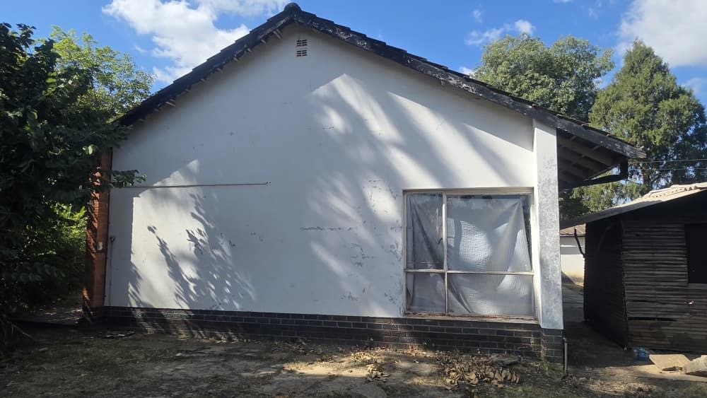 Property photo 2 of 12 White Single-Story House With A Dark Tiled Gable Roof, A Large Window With A Sheer Curtain On The Right, And A Brick Base, Shaded By Nearby Trees.